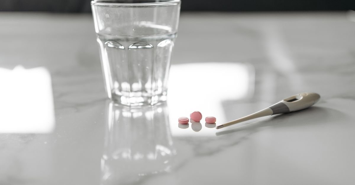 Glass of water, a thermometer and a few tablets on a pale surface during flu illness