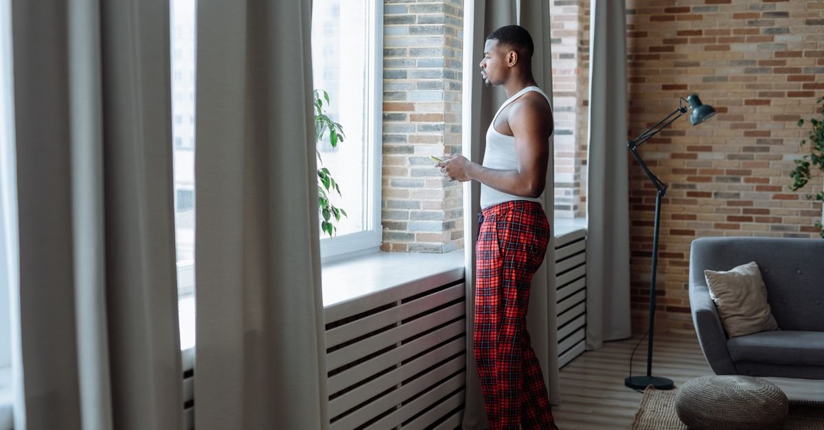 A man in pyjamas standing at a living room window in the middle of the morning