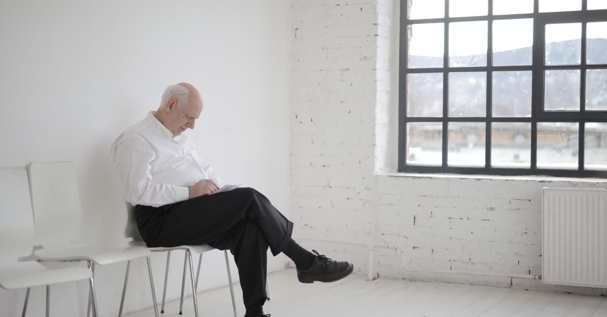 Older man sitting alone on a chair by a tall window, waiting for brain fog test results